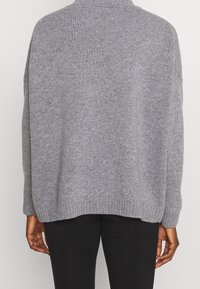 Grijze oversized trui gemaakt van zacht breimateriaal. Heeft een hoge halslijn en een geribbelde zoom. Van achteraf weergegeven, met lange mouwen.