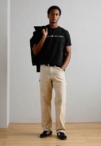 Zwarte t-shirt met witte tekst, beige broek, zwarte schoenen met witte strepen en een zwarte jas over een schouder.