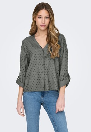 Dark green button-up blouse with a V-neck, textured polka dot pattern, and rolled sleeves, complemented by a relaxed fit.