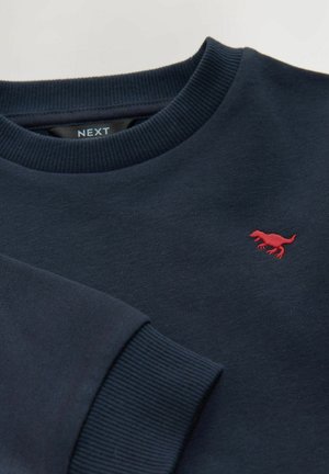 Temno modra športna majica, mešanica bombaža, z ribastim izrezom in manšetami, z majhnim rdečim vezenim logotipom dinozavra na prsih.