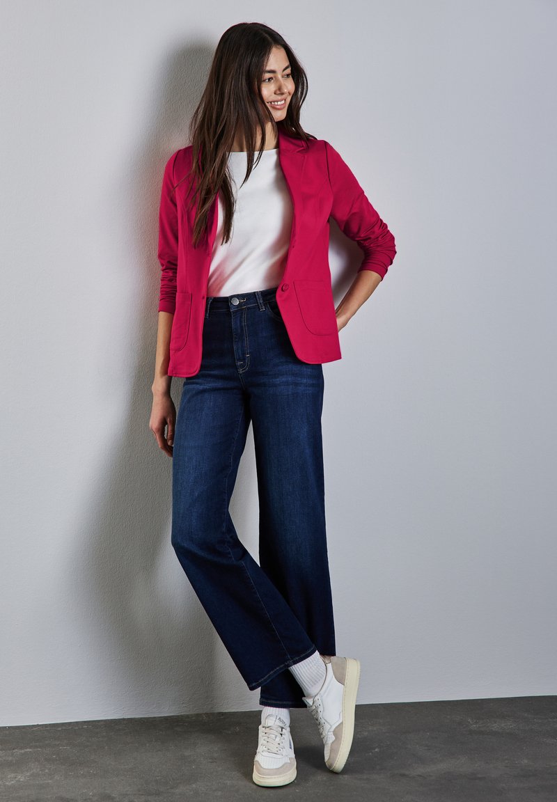 Fuchsia-Blazer, weißes Hemd, dunkelblaue weite Jeans und cremefarbene Sneakers. Der Blazer hat zwei Taschen und einen Knopf. Glatte Texturen.
