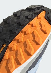Semelle de chaussure de trail running avec une semelle extérieure en caoutchouc noire et orange, des crampons profonds, des détails texturés et une semelle intermédiaire blanche pour le soutien.