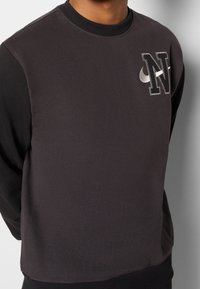 Mörkgrå sweatshirt med långa ärmar, ribbad krage och en texturerad logotyp med "N" och en böjd accent i ljusare grått.