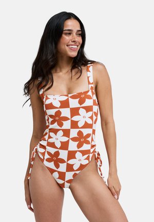Kobieta uśmiechająca się, ubrana w jednoczęściowy kostium kąpielowy w pomarańczowo-biały w kratkę z kwiatowym wzorem i wiązaniami po bokach na jednolitym tle.