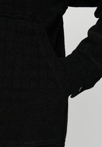 Μαύρο υφασμάτινο μπουφάν με υφή, μανίκι με το χέρι να έχει εισαχθεί μερικώς σε ένα πλαϊνό τσέπη, με μια μικρή μαύρη κουμπί στον καρπό.