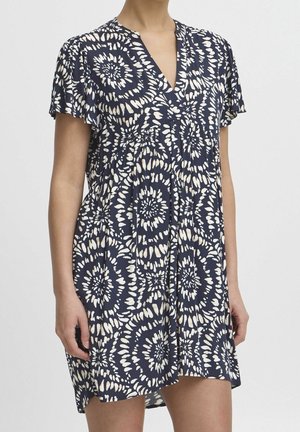 Woman wearing a navy blue short-sleeve dress with white circular tie-dye patterns and a V-neckline, standing against a plain background.