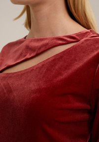 Kraljevsko crvena baršunasta majica s širokim izrezom i asimetričnim izrezom. Mekana tekstura i glatka završna obrada ističu područje ramena.
