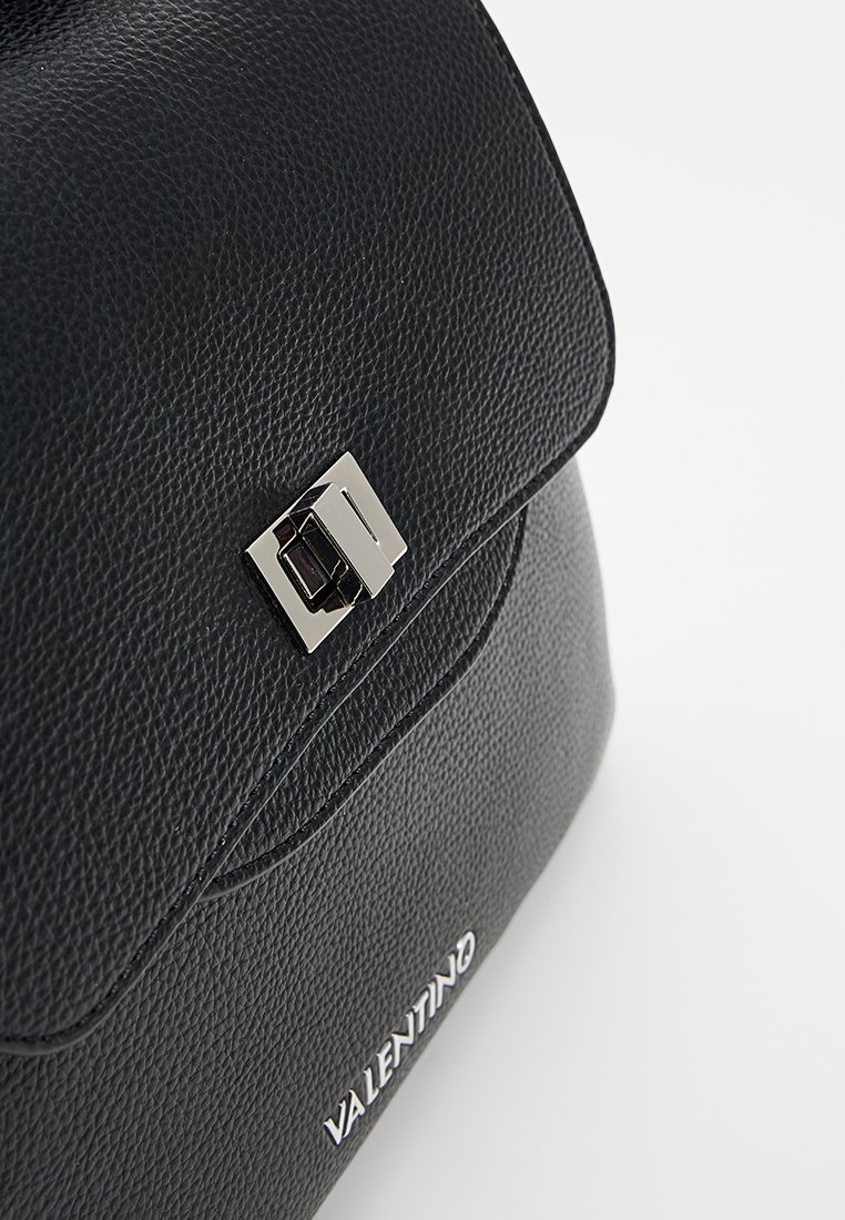 Sac à main en cuir noir avec une surface texturée, un rabat supérieur, des accessoires en métal argenté, et un logo affiché en bas.