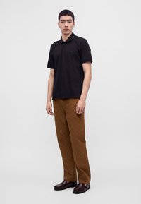 Jeune homme debout portant un polo noir à manches courtes, un pantalon marron à motifs et des mocassins marron foncé sur un fond gris uni.