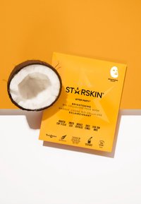 Kokosnootschulp naast een felgele verpakking van het STARSKIN After Party bio-cellulose gezichts-maske, met tekst die voordelen en ingrediënten benadrukt.