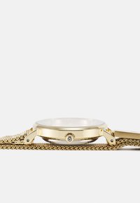 Montre en métal doré avec un cadran rond et transparent et une lunette lisse. Elle présente un bracelet en maille texturée et une petite pierre décorative sur la couronne.