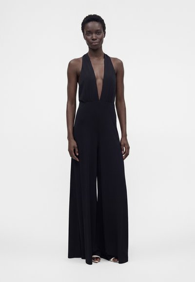 Femme portant une combinaison noire sans manches avec un décolleté en V profond et un pantalon à jambes larges, debout devant un fond clair uni.