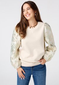 Sweatshirt crème avec des manches texturées à fleurs pastel vert et or. Doté d'un col rond et d'une coupe décontractée. Associé à un jean bleu.