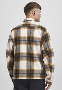 Plaid flannel shirt in white, gray, and brown with yellow accents. Features a collar and buttoned cuffs, showcasing a soft texture.