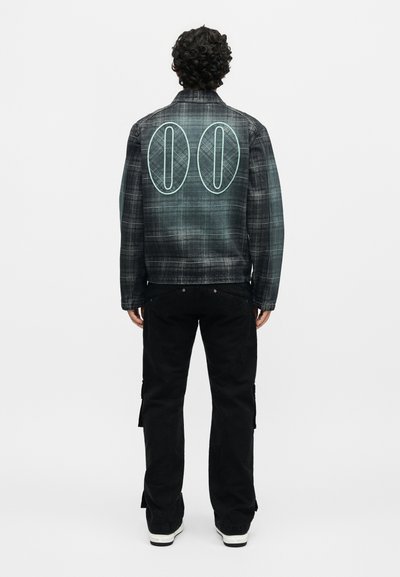 Άνδρας με σγουρά μαλλιά που φοράει σκούρο καρό μπουφάν με μεγάλο "00" στην πλάτη, μαύρο παντελόνι και παπούτσια με λευκή σόλα, όρθιος με την πλάτη προς τα εμπρός.
