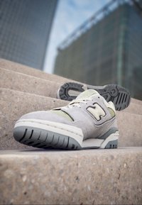 Chaussure de sport grise et beige en textile et caoutchouc, avec un bout arrondi, des coutures blanches et une semelle texturée avec des accents.