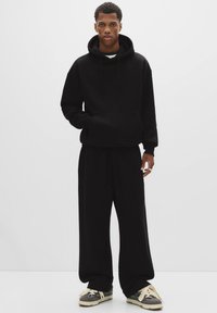 Sort hættetrøje med en frontlomme og snøre i huen, parret med matchende løstsiddende sweatpants. Sorte og grå sneakers med beige accents.