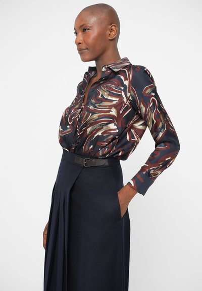 Langærmet bluse med mørkeblå baggrund, abstrakte flerfarvede mønstre i nuancer af burgunder, grøn og creme; sat sammen med en marineblå nederdel.