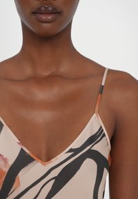 Débardeur en tissu léger beige avec des motifs abstraits noirs et oranges. Il a des bretelles fines et un décolleté en V profond. Texture douce.