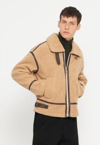 Beige fleecejakke med brune læderdetaljer, lynlåslukning, stor krave og to forsøgspokker, der har et struktureret, hyggeligt udseende.