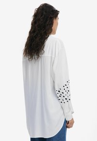 Weißes Langarmshirt mit schwarzen Punktmustern auf den Ärmeln, weicher Stoff, lockere Passform und abgerundetem Saum.