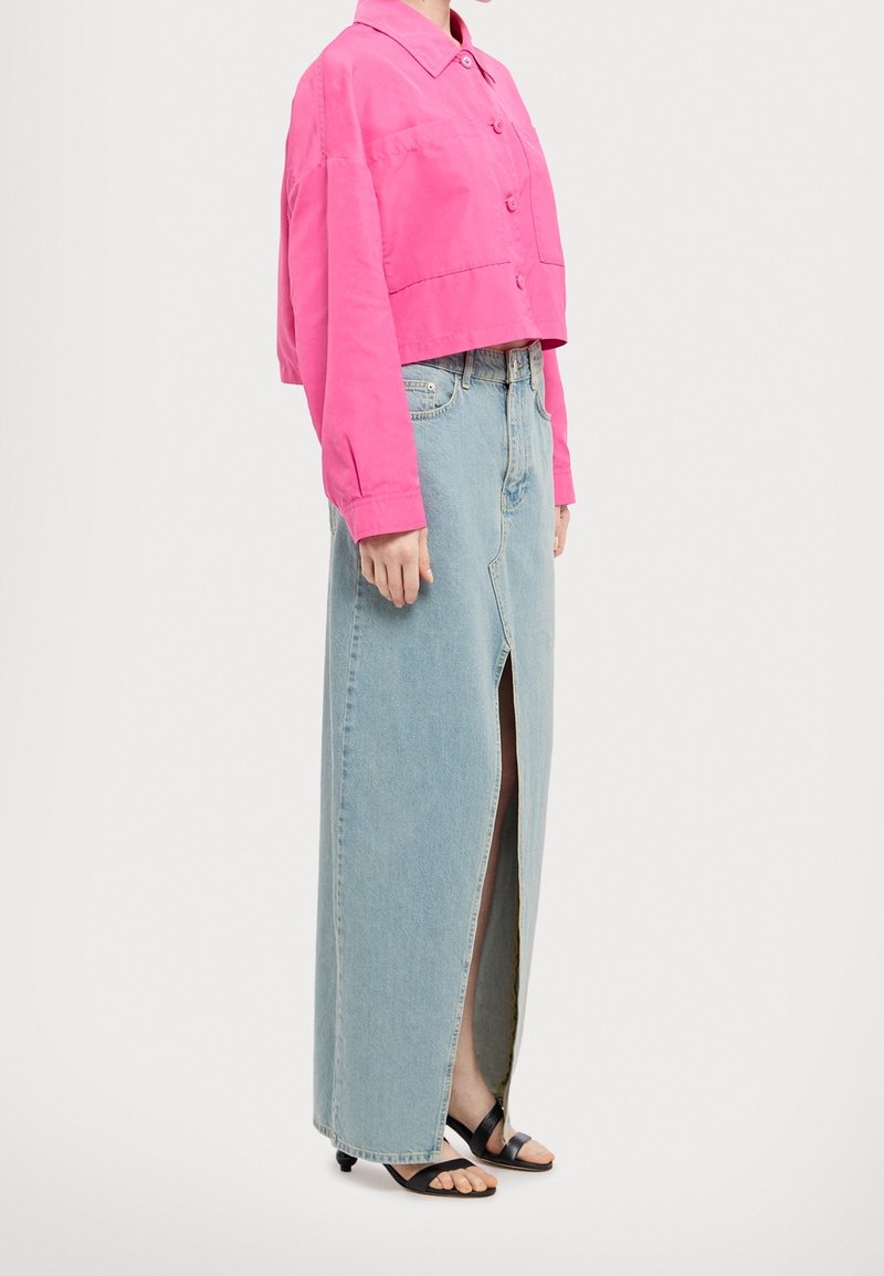 Mujer con una camisa corta de botones de color rosa brillante, falda larga de mezclilla azul clara de talle alto con abertura frontal y sandalias negras de punta abierta.