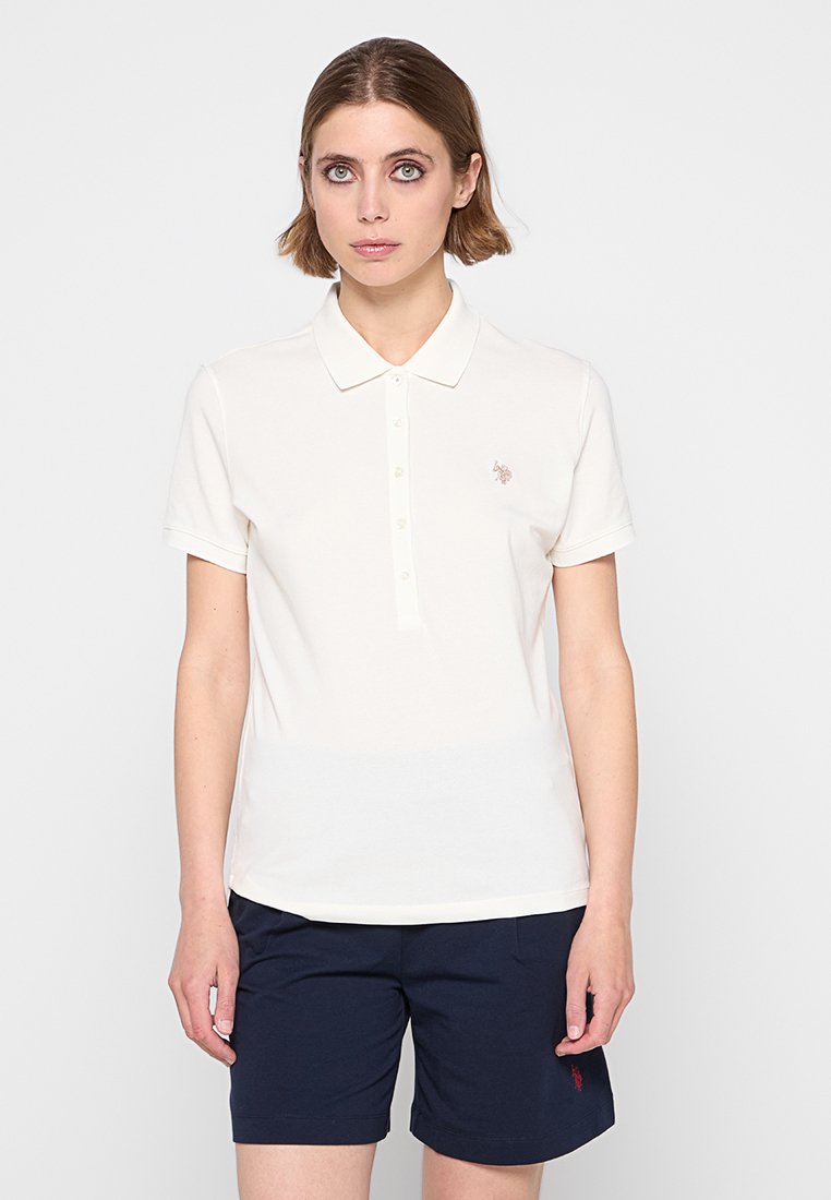 Polo shirt crema con maniche corte e apertura con bottoni, con un piccolo logo ricamato sul petto. Abbinato a pantaloni corti blu navy.