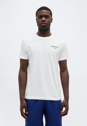 Άνδρας με κοντά μαλλιά που φοράει λευκό μπλουζάκι Björn Borg και μπλε παντελόνι, στέκεται μπροστά σε μια απλή λευκή επιφάνεια.