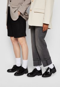 Due persone indossano un blazer beige e uno grigio, pantaloni corti neri, jeans grigi e scarpe lucide nere con chiusura a lacci, completati da calzini bianchi.