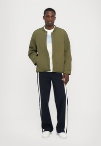 Jachetă matlasată de culoare verde măslin cu guler rotund, purtată peste o cămașă albă cu dungi albastre. Asortată cu pantaloni bleumarin cu dungi albe pe lateral.