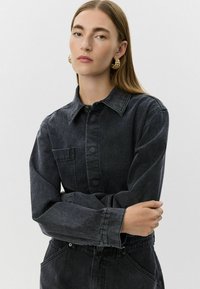 Sötét mosású farmering, hegyes gallérral, elülső zsebekkel és gombos záródással. Lazább ujjakkal és texturált anyaggal rendelkezik.