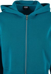 Teal zip-up dukserica napravljena od mekanog materijala s dugim rukavima, kapuljačom i rebrastim manžetama. Ima srebrni zatvarač i Urban Classics oznaku unutra.