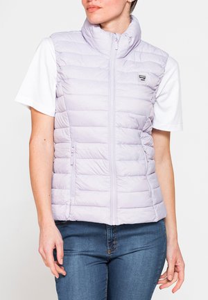 Donna che indossa un gilet trapuntato senza maniche viola chiaro con zip frontale sopra una maglietta bianca a maniche corte e jeans blu, in piedi davanti a uno sfondo bianco.