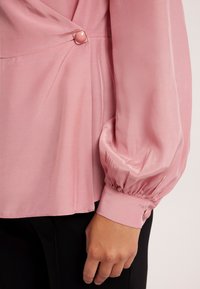 Różowa bluzka o gładkiej satynowej teksturze, z fasonem typu wrap, bufiastymi rękawami i ozdobnym guzikiem z boku.