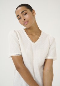 Blouse blanche à manches courtes avec un col en V, en tissu texturé présentant de petits points en relief, et des accents en dentelle festonnée au niveau du col et des manches.