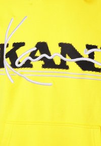 Sudadera amarilla con un logo negro impreso, que presenta acentos bordados en blanco y un acabado texturizado. Incluye un bolsillo frontal.