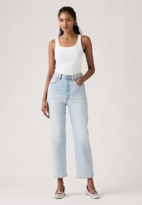 Γυναίκα που στέκεται με το ένα χέρι στην τσέπη ανοιχτόχρωμων τζιν ψηλόμεσων, φορώντας λευκό τοπ χωρίς μανίκια και ασημένιες μπαλαρίνες, σε απλό φόντο.