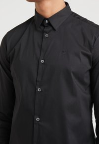 Camisa negra con cuello clásico, manga larga y un diseño ajustado. Presenta botones negros y un logo bordado sutil en el pecho.