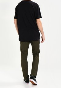 T-shirt nera a maniche corte, vestibilità ampia, abbinata a pantaloni verdi oliva aderenti e sneakers nere con dettagli bianchi, vista da dietro.