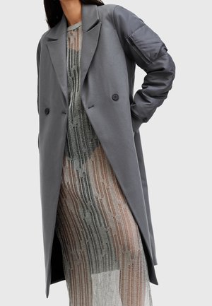 Long manteau gris avec poche matelassée sur la manche, porté sur une robe en chaîne argentée transparente, sur une personne avec la main sur la hanche.