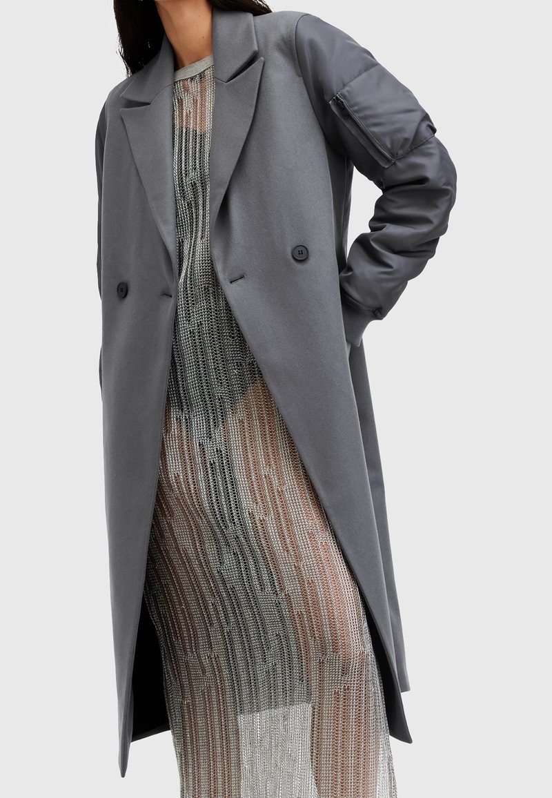 Long manteau gris avec poche matelassée sur la manche, porté sur une robe en chaîne argentée transparente, sur une personne avec la main sur la hanche.