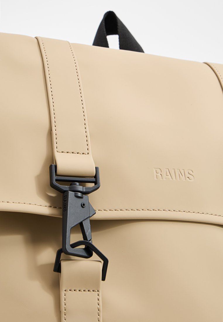 Zaino impermeabile beige realizzato in materiale liscio, con hardware nero a contrasto e una chiusura con fibbia distintiva. Logo "RAINS" visibile.
