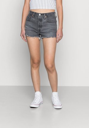 Femme portant un short en jean noir taille haute avec des bords effilochés, un crop top blanc sans manches avec des rayures verticales noires, et des baskets montantes blanches.