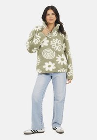 Pullover verde fluffy con motivi floreali bianchi e spirali. Presenta un colletto alto e maniche lunghe, abbinato a jeans azzurri chiari e sneakers.