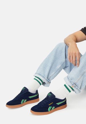 Mornarsko plave tenisice od brušene kože sa zelenim i žutim detaljima, s gumenim potplatom boje tabaka. Uparene s laganim trapericama i bijelim čarapama sa zelenim prugama.