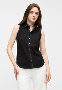 Νεαρή γυναίκα με μακριά καστανά μαλλιά, ντυμένη με αμάνικο μαύρο πουκάμισο με κουμπιά και λευκό παντελόνι, στέκεται μπροστά σε απλό φωτεινό φόντο.