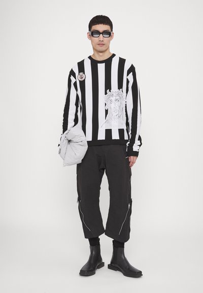 Gestreifter Schwarz-Weiß-Pullover mit Grafikdruck, schwarze Cargo-Hosen mit Reißverschlüssen und schwarze Stiefeletten. Modell hält eine graue Jacke.