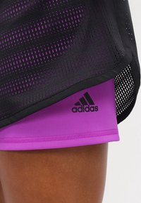 Schwarzes Mesh-Sporttop, das über lila Kompressionsshorts getragen wird, mit einem geprägten schwarzen Adidas-Logo, weicher Textur und atmungsaktivem Design.