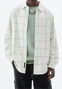 Camisa beige clara y ligera con cuello, botones delanteros completos y tejido texturizado. Lleva una capa sobre un suéter verde menta. Se ven pantalones negros.