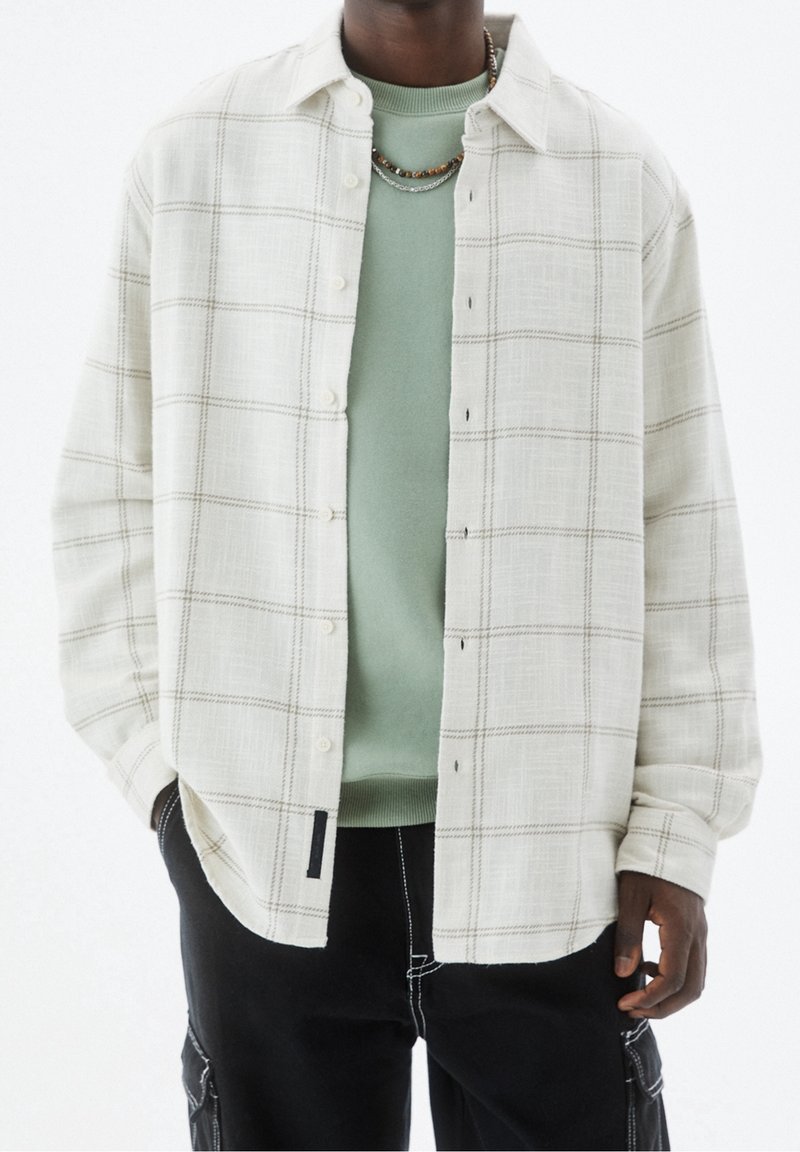 Camisa beige clara y ligera con cuello, botones delanteros completos y tejido texturizado. Lleva una capa sobre un suéter verde menta. Se ven pantalones negros.